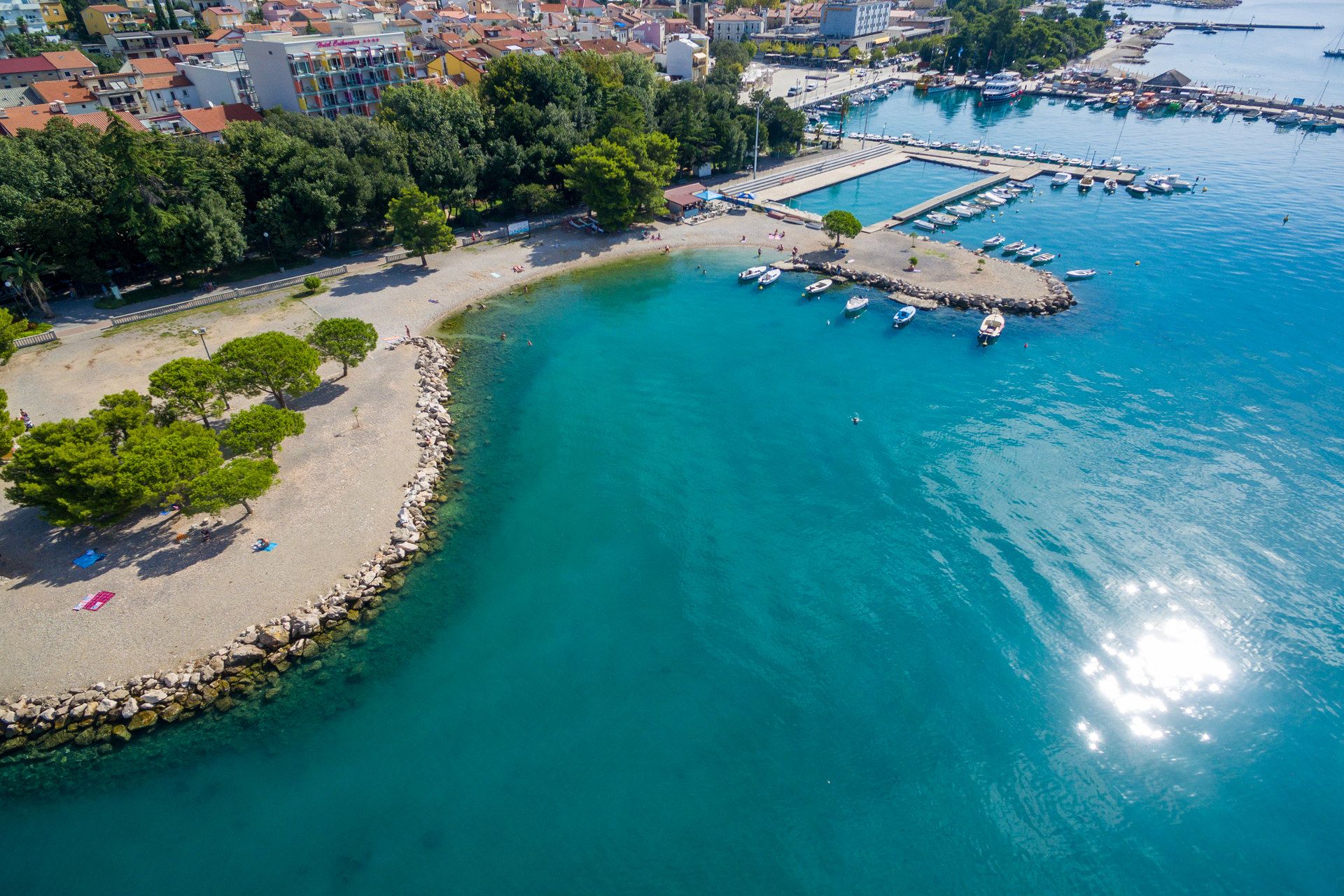 A pebble beach curving round a spit with pine trees, clear green water, small boats and the harbour behind