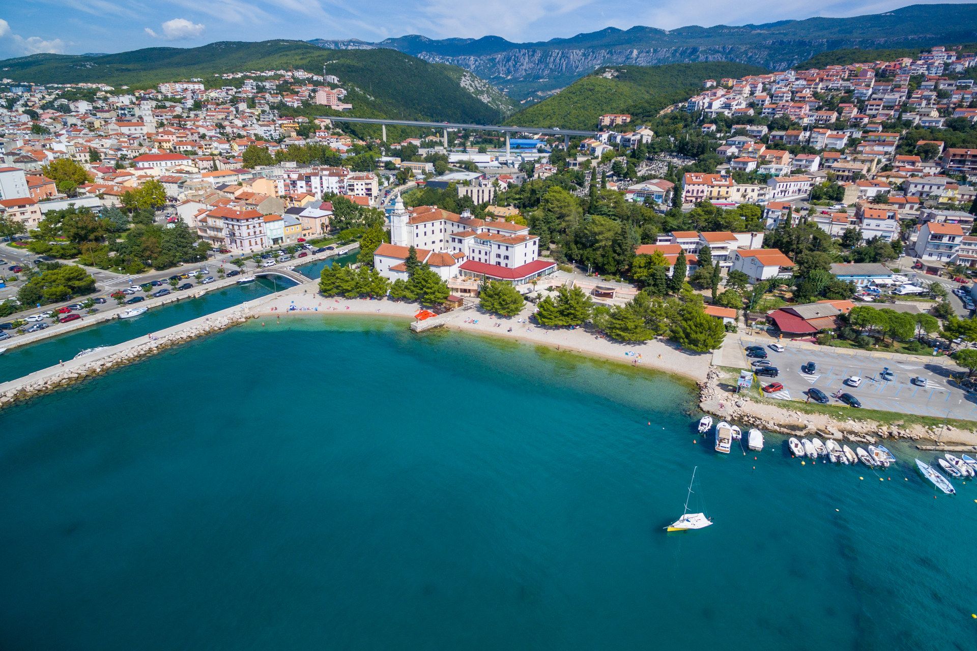 Aerial view of a pebble beach in front of a white hotel with a bell tower, a river mouth and a footbridge to the left, the town climbing green hills behind, a road viaduct between them