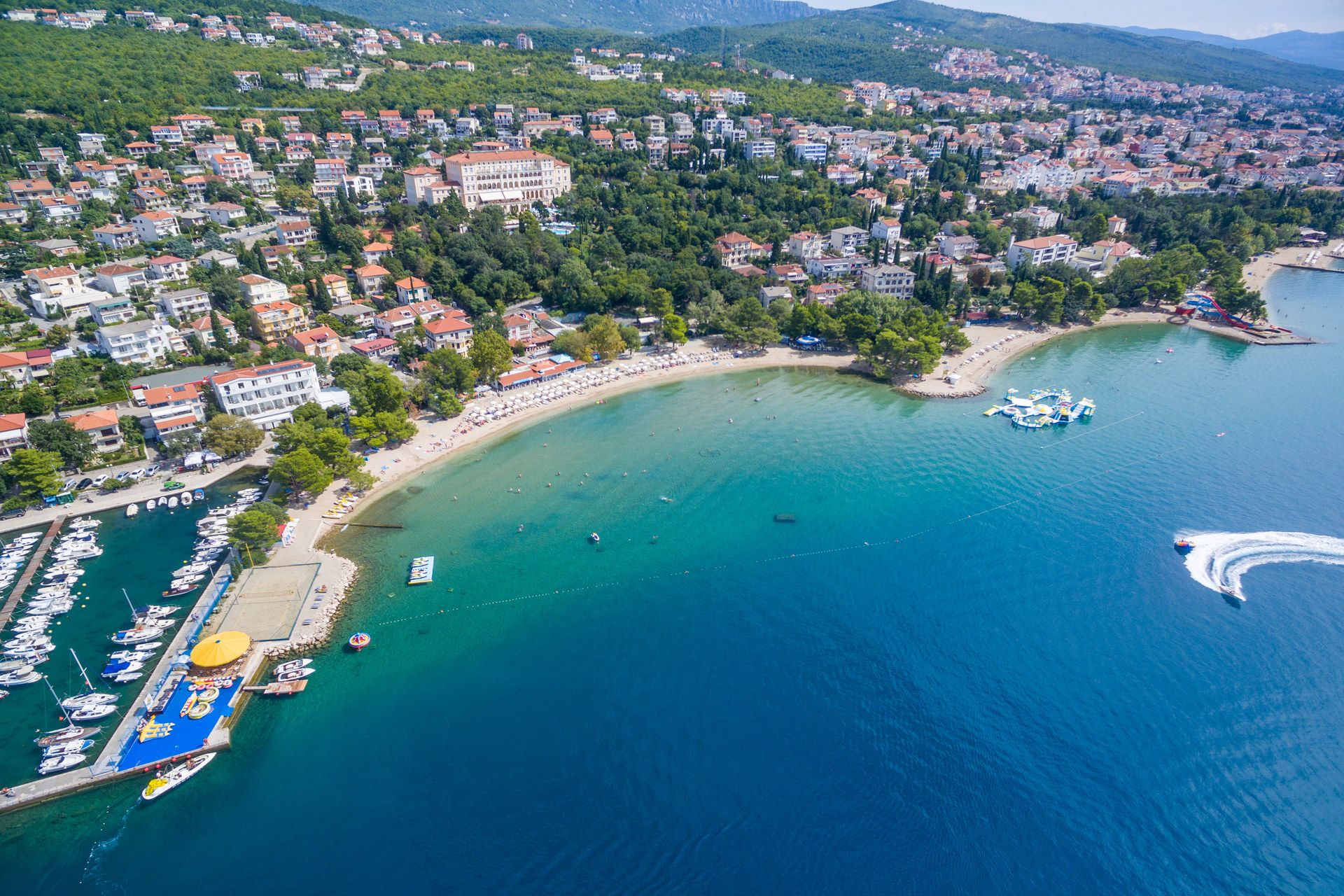 Aerial view of a long curved shore: a small harbour, a beach lined with parasols, a point with pine trees, then a second bay with an inflatable water park and a slide, the town on the hill above