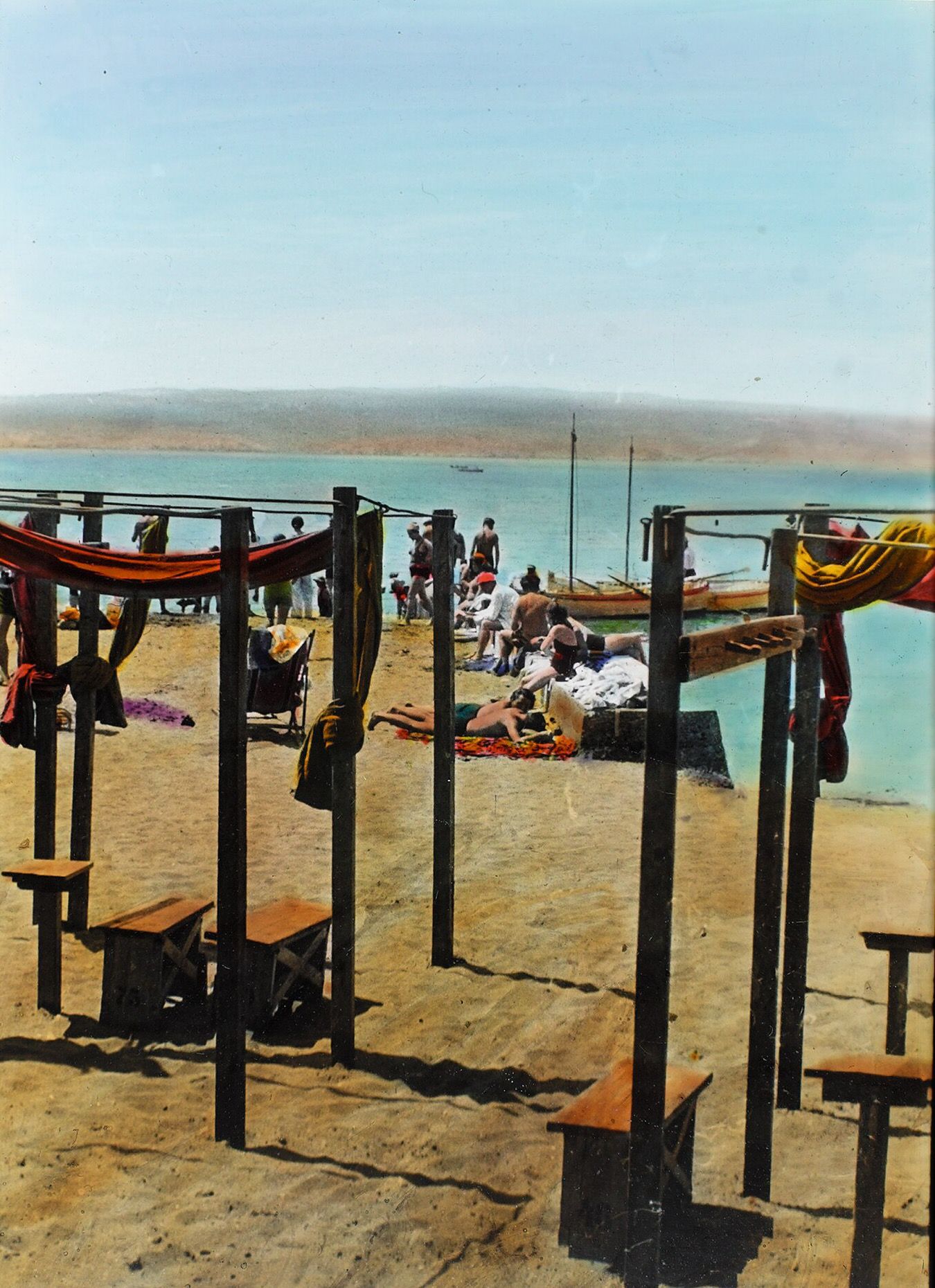 Hand-coloured photograph from 1936 of a sandy beach with wooden changing frames, bathers and two sailing boats