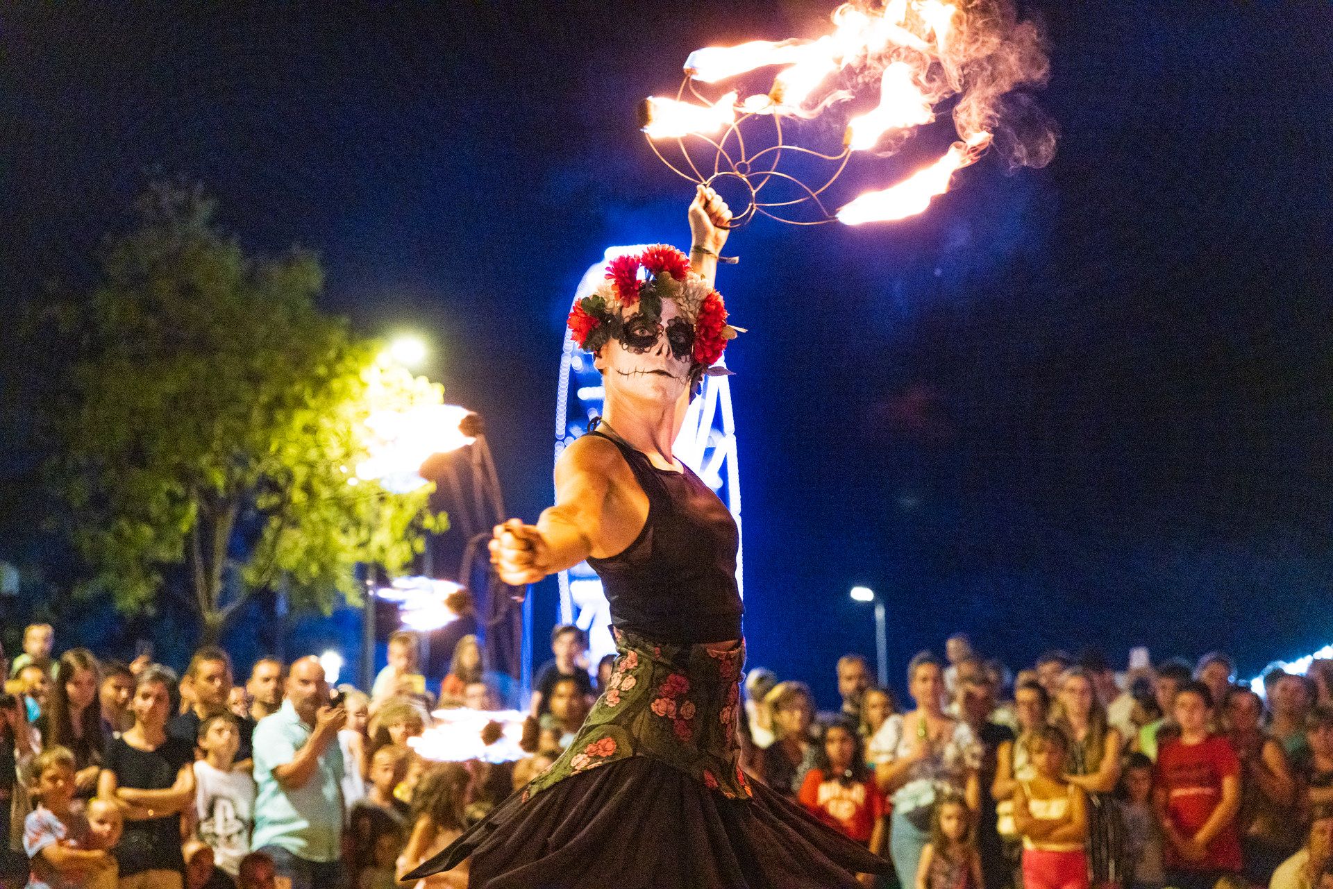 A fire performer in skull make-up and a crown of red flowers swings burning torches at night, a crowd watching behind her