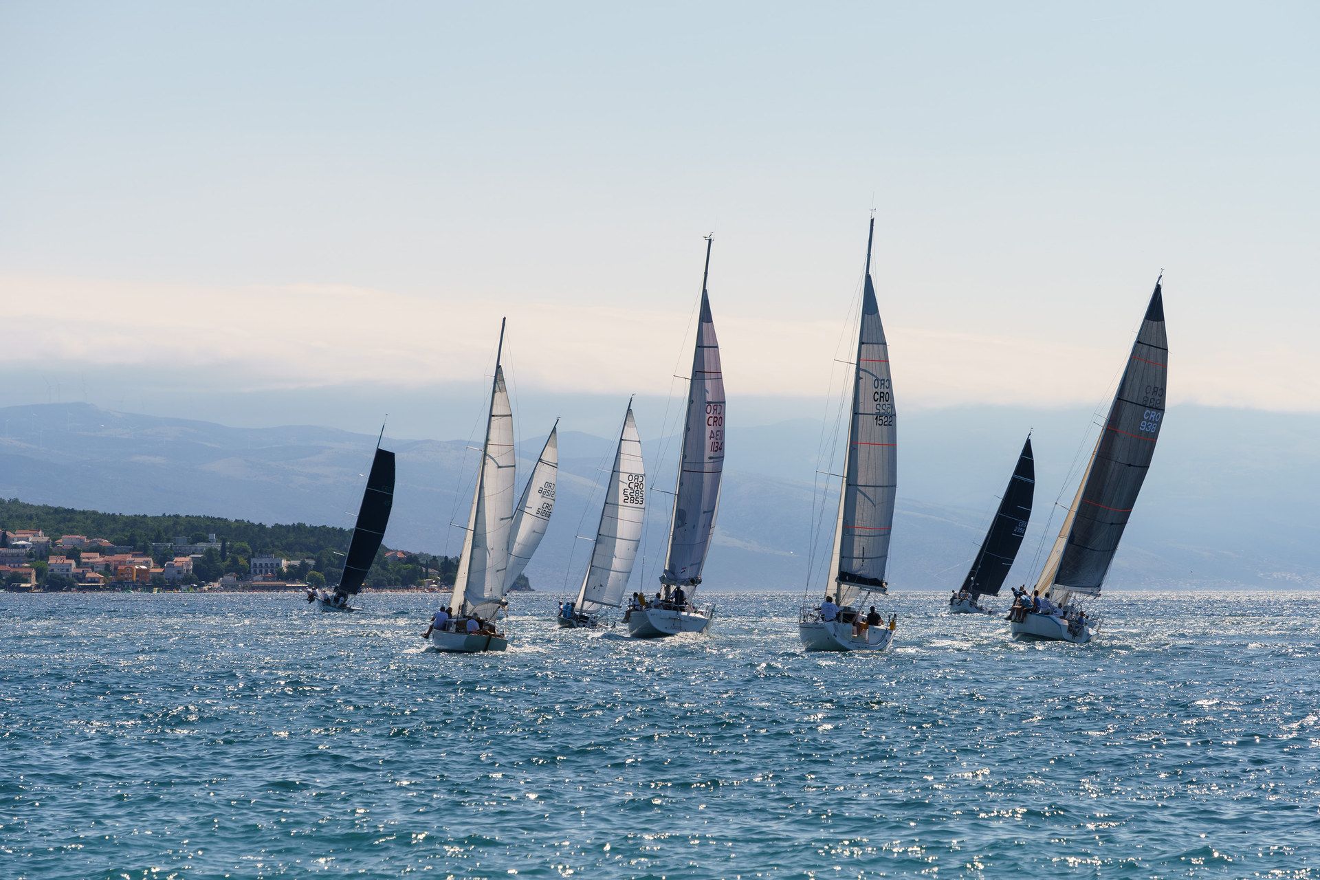 Seven sailing yachts racing close together on a bright sea, the coast and hills behind