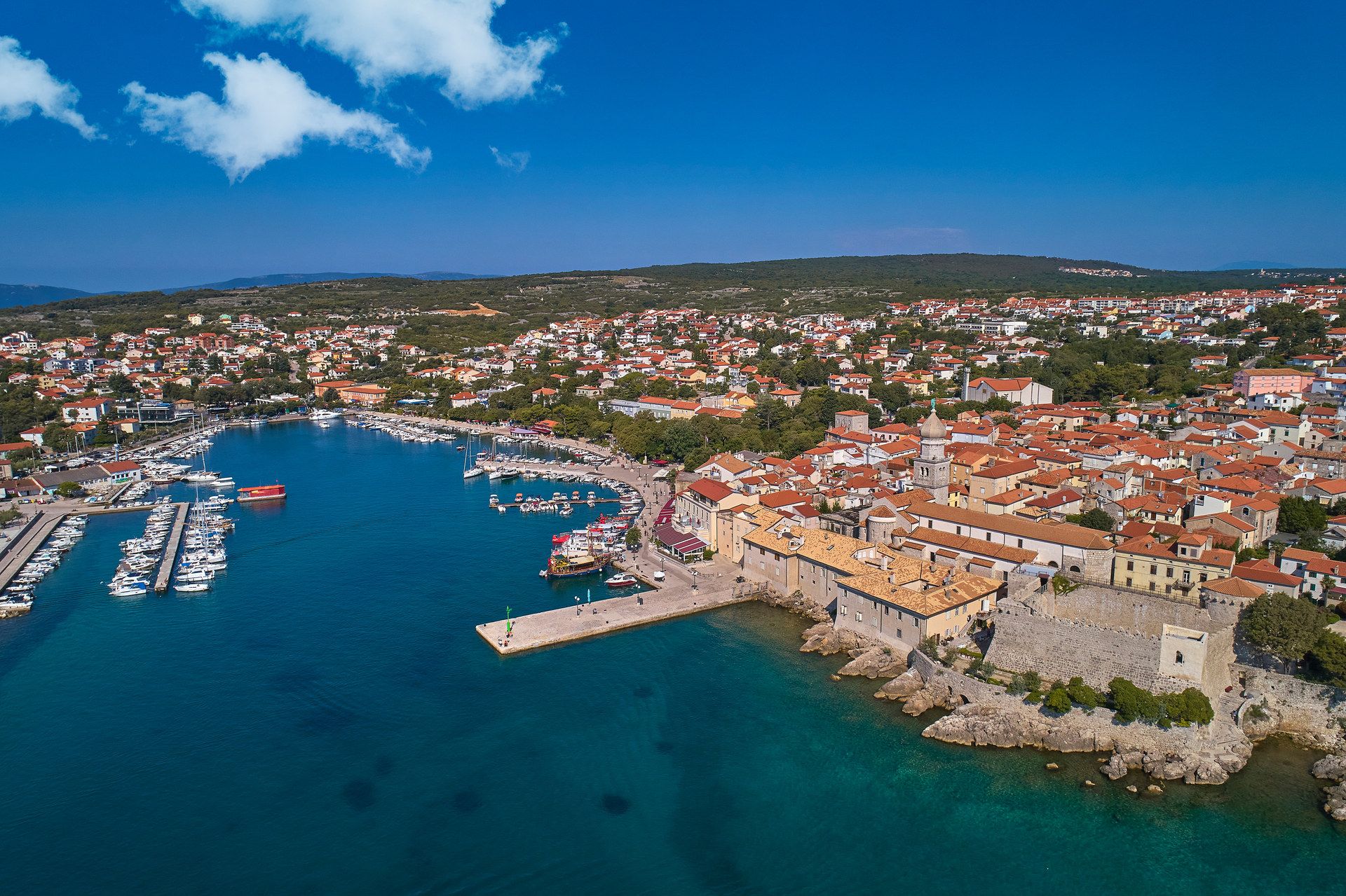 Aerial view of Krk: red roofs and a bell tower inside stone walls on the shore, a harbour full of boats to the left