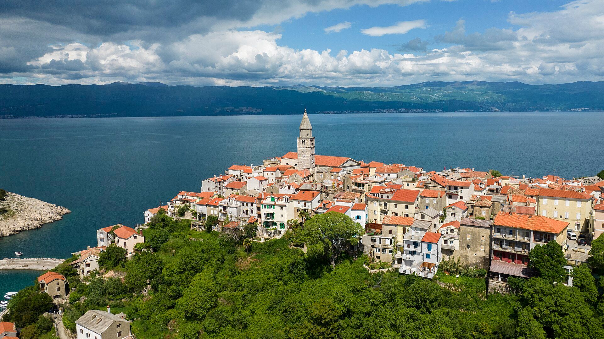 Aerial view of Vrbnik: stone houses and a bell tower on a cliff above the sea, the mainland in the distance